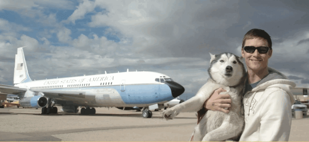 A smiling man wearing sunglasses and a light hoodie holds a relaxed Siberian Husky in his arms. Behind them is a large aircraft painted with "United States of America," parked on an airfield under a dramatic, cloudy sky.