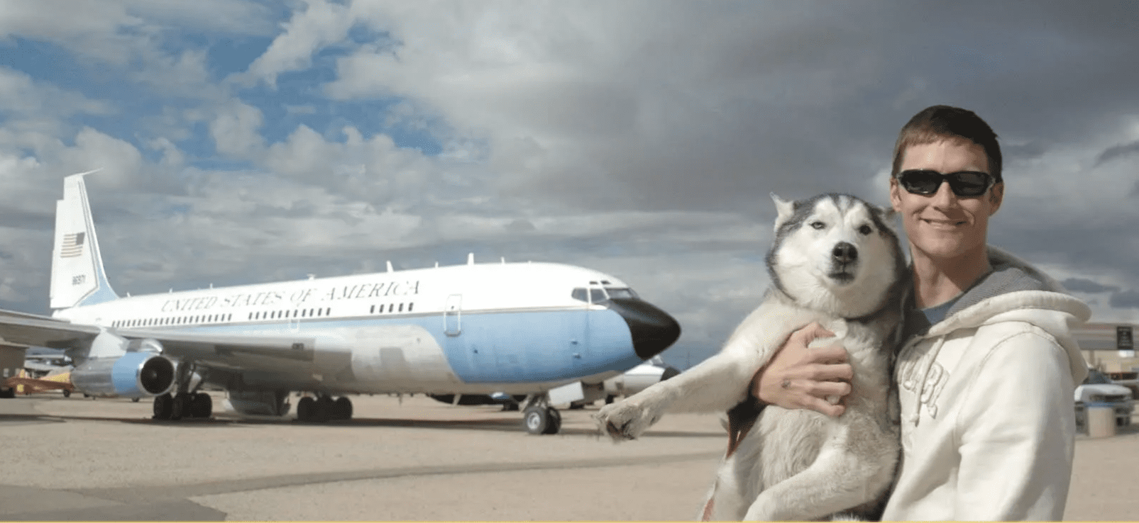 A smiling man wearing sunglasses and a light hoodie holds a relaxed Siberian Husky in his arms. Behind them is a large aircraft painted with "United States of America," parked on an airfield under a dramatic, cloudy sky.